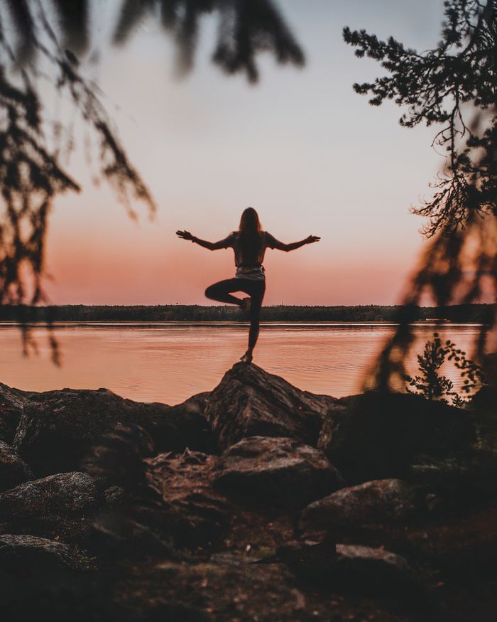 service-02 Silhouette of a woman practicing yoga on rocks by the water at sunset, creating a serene outdoor scene.